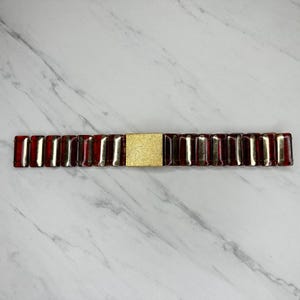 May include: A red and gold bracelet with a rectangular gold clasp. The bracelet is made of rectangular red and gold segments. The clasp is a solid gold color and is rectangular in shape.