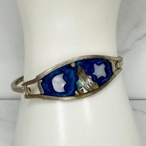 May include: Silver-tone bracelet with a blue enamel inlay and iridescent abalone shell accents. The bracelet features two white, crescent-shaped inlays and a hinged design for easy wear. A unique piece of jewelry.