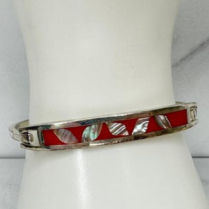 May include: Silver-tone hinged bangle bracelet with a red enamel inlay. The bracelet features iridescent shell accents and a rectangular design. The bracelet is shown on a white mannequin arm.