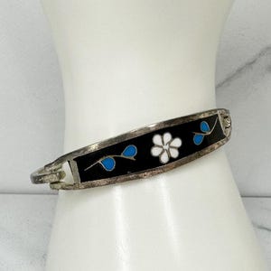 Peut inclure: Bracelet articulé argenté avec un centre en émail noir. Le bracelet présente des motifs de fleurs blanches et de feuilles bleues. Le bracelet est présenté sur une surface blanche.