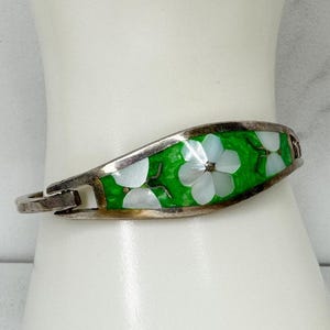 May include: Silver-tone hinged bracelet with a vibrant green enamel inlay. The inlay features white flower designs with mother-of-pearl petals. The bracelet is a hinged style, designed to be worn on the wrist.