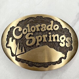 May include: Oval-shaped brass belt buckle with the words "Colorado Springs" in a stylized font. The buckle features a mountain range and decorative scrollwork. The buckle has a vintage look.