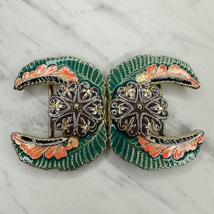 Könnte beinhalten: Eine verzierte, zweiteilige Schnalle mit floralem Design. Die Schnalle hat ein grünes, orangefarbenes und schwarzes Blättermuster mit einer zentralen braunen und silbernen Blume. Das Metall ist goldfarben.