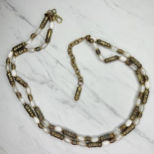 May include: A vintage belt featuring a double strand design. The belt is composed of white, oval-shaped beads, interspersed with gold-toned, cylindrical and patterned beads. A gold-toned chain and clasp complete the belt, which is displayed on a marble surface.