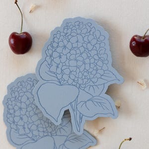 Puede incluir: Dos recortes de papel azul claro con forma de hortensia con arte lineal floral detallado. Cerezas frescas y maduras y pequeños pétalos blancos dispersos, todo sobre un fondo blanco roto. El diseño es delicado e inspirado en la naturaleza.