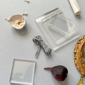 May include: A flat lay featuring a variety of objects, including two clear glass squares, a small white ceramic bowl with dried white flowers, a grey fabric ribbon tied in a bow, a rolled up piece of white paper, two purple figs, and a gold framed mirror.