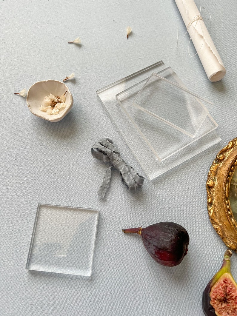 May include: A flat lay featuring a variety of objects, including two clear glass squares, a small white ceramic bowl with dried white flowers, a grey fabric ribbon tied in a bow, a rolled up piece of white paper, two purple figs, and a gold framed mirror.