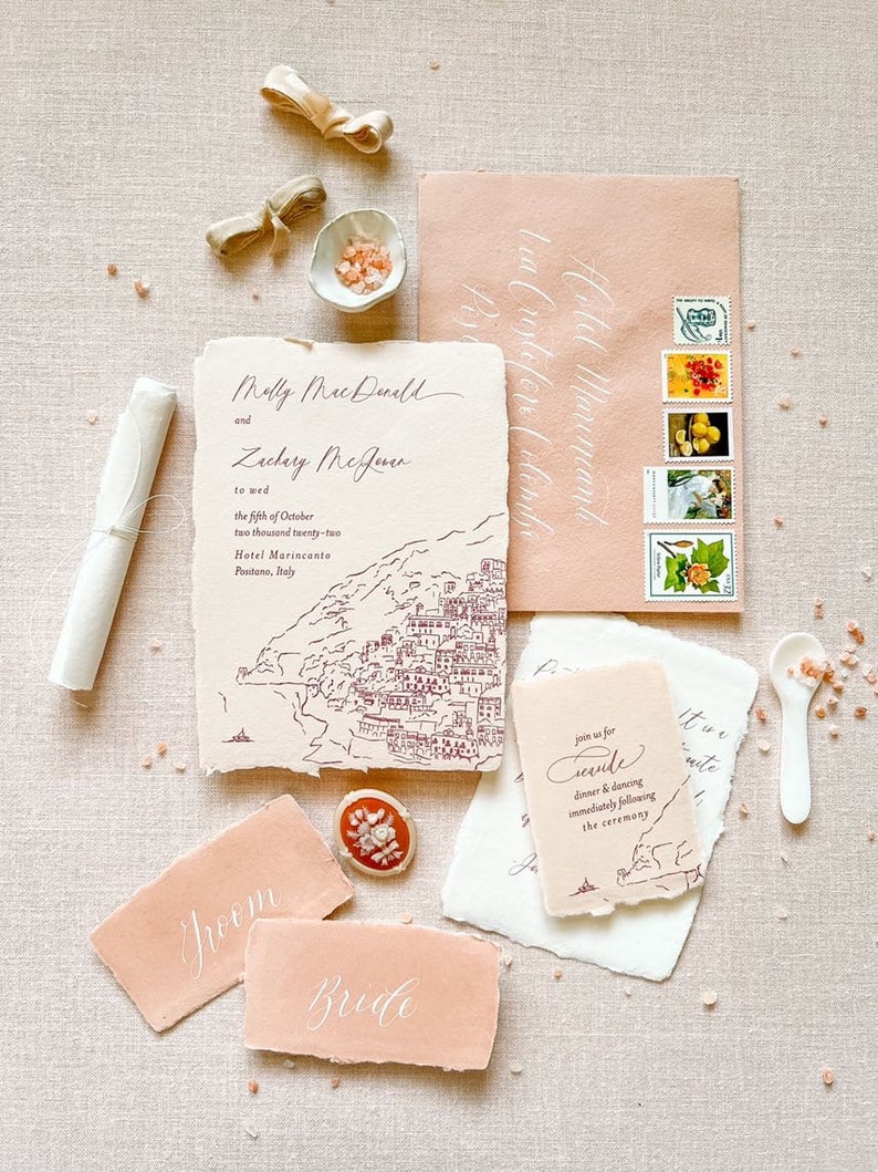 May include: Wedding invitation suite with a hand-drawn illustration of a coastal town. The invitation is on textured paper with a peach envelope. The invitation reads "Melly MacDonald and Zachary McJener to wed the fifth of October two thousand twenty-two Hotel Marincanto Positano, Italy". The RSVP card reads "Join us for seconde dinner and dancing immediately following the ceremony".