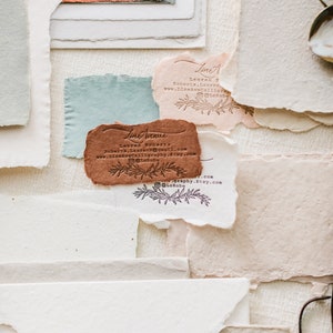 May include: An assortment of handmade paper pieces in various colors and textures. The image features torn-edge paper in shades of white, peach, blue, and brown. Several pieces have printed text and floral designs. A vintage-style metal object is visible in the upper right corner.