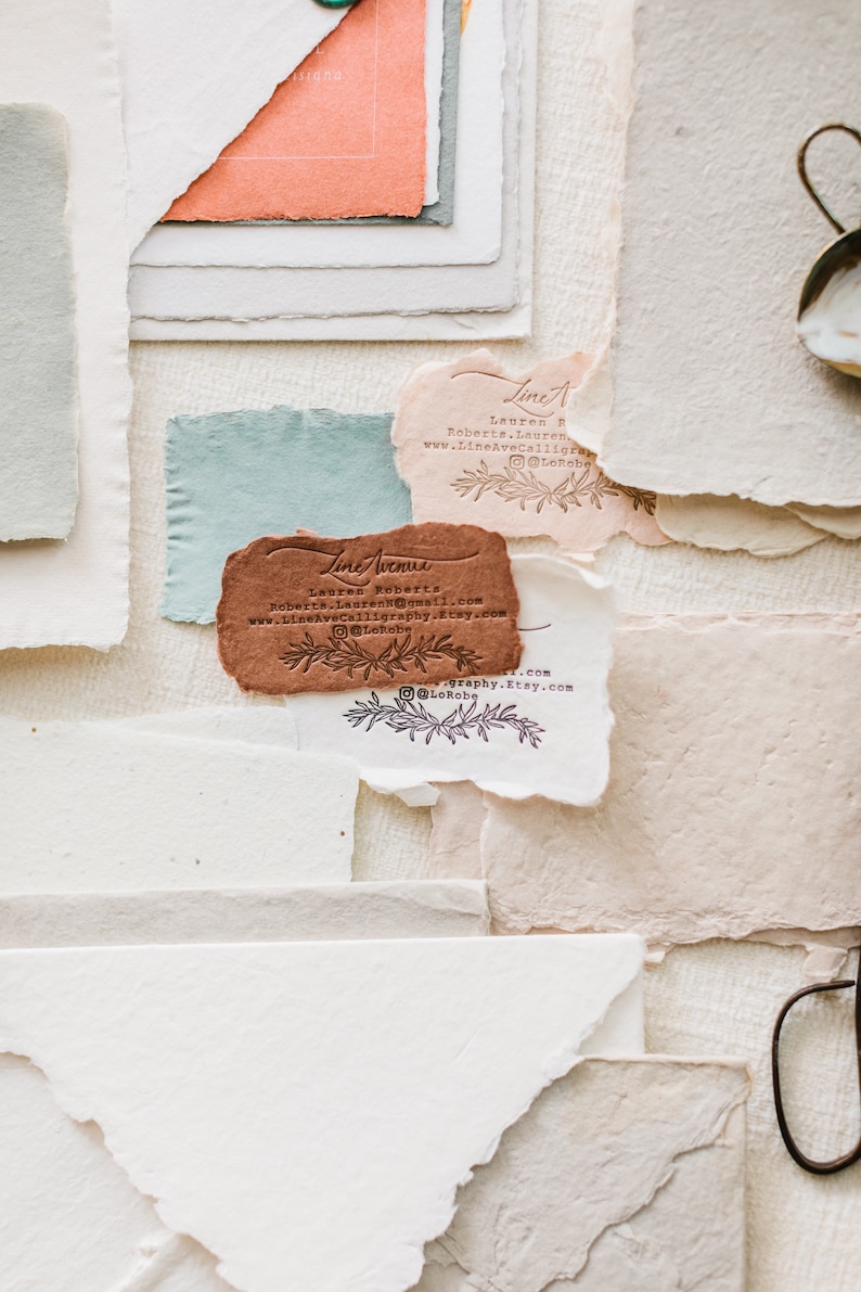 May include: An assortment of handmade paper pieces in various colors and textures. The image features torn-edge paper in shades of white, peach, blue, and brown. Several pieces have printed text and floral designs. A vintage-style metal object is visible in the upper right corner.