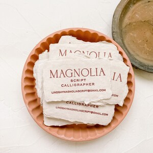 May include: A stack of off-white business cards with the name "MAGNOLIA" in burgundy script, along with "SCRIPT CALLIGRAPHER" and an email address. The cards rest in a peach-colored, scalloped-edge dish. A weathered, brown dish is in the background.
