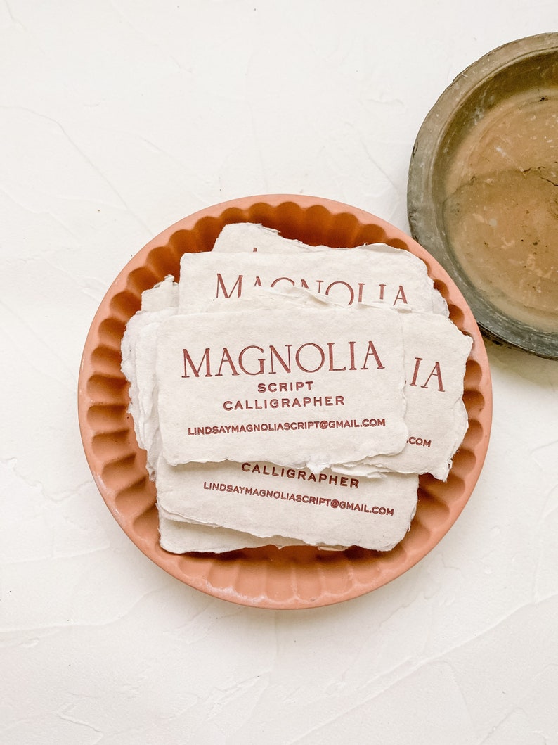 May include: A stack of off-white business cards with the name "MAGNOLIA" in burgundy script, along with "SCRIPT CALLIGRAPHER" and an email address. The cards rest in a peach-colored, scalloped-edge dish. A weathered, brown dish is in the background.
