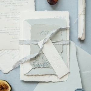 May include: A stack of wedding invitations with a blue and white color scheme. The invitations are tied together with a white ribbon and have a delicate floral design. The invitations are on a light blue background with a blue wax seal.