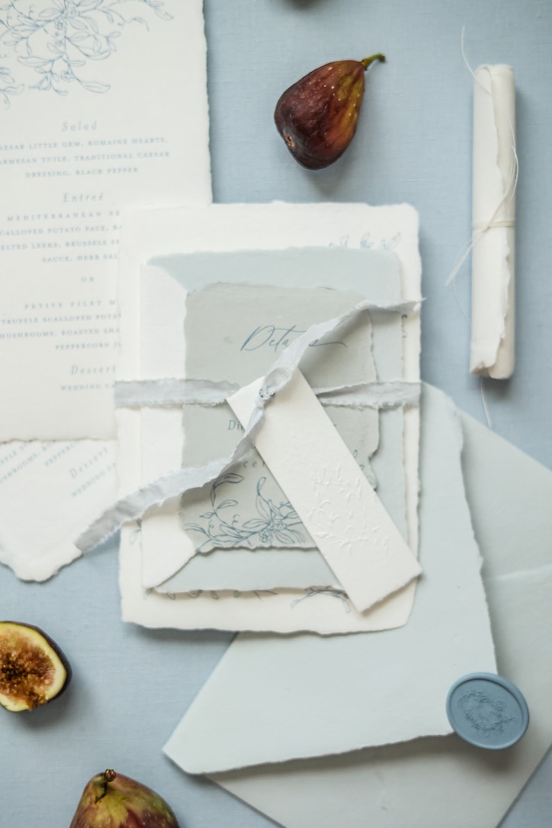 May include: A stack of wedding invitations with a blue and white color scheme. The invitations are tied together with a white ribbon and have a delicate floral design. The invitations are on a light blue background with a blue wax seal.