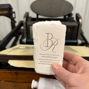 May include: A stack of off-white business cards with the embossed initials "BR" and the text "Brittany Roberts Exclusive Timeless Wedding Imagery". The cards are held in front of an antique printing press and a tube of white paint.