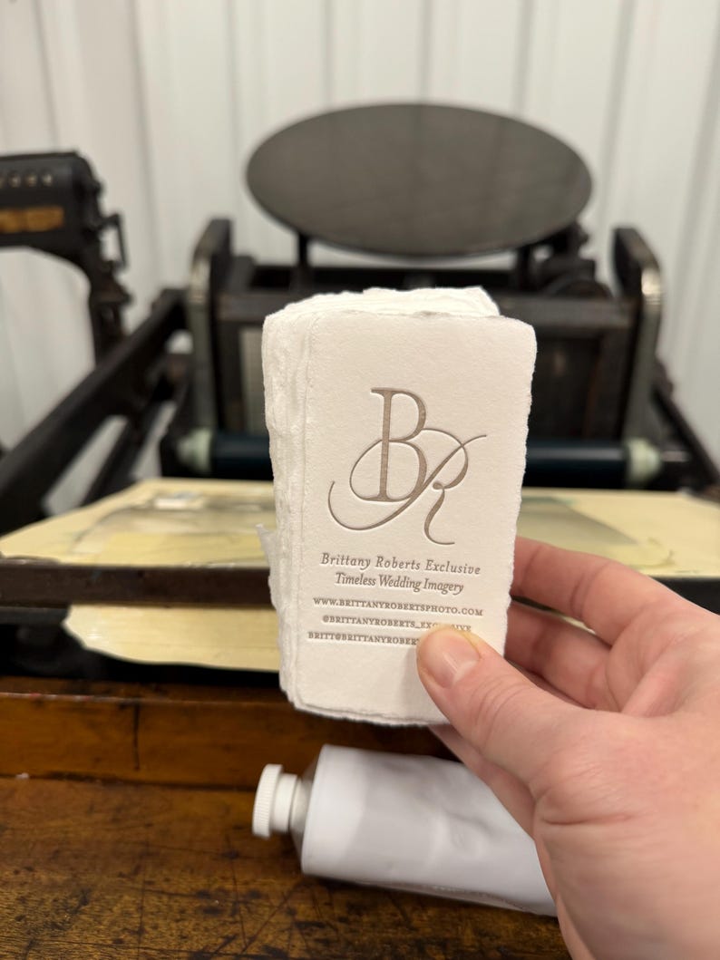 May include: A stack of off-white business cards with the embossed initials "BR" and the text "Brittany Roberts Exclusive Timeless Wedding Imagery". The cards are held in front of an antique printing press and a tube of white paint.