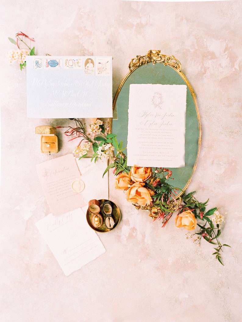 May include: Wedding invitation suite with a gold-framed mirror, a white envelope with stamps, a pink and white invitation card, and a small box with a ring. The invitation card has calligraphy text that reads "Tyler and Jordan" and the date of the wedding. The background is a light pink.