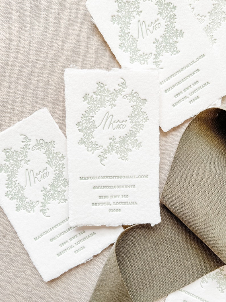 May include: Off-white business cards with a torn edge, featuring a floral wreath design and the text "Mery 160." Additional text includes contact information. A muted green ribbon is visible in the background.