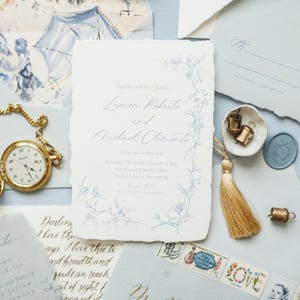 May include: A wedding invitation suite with a delicate floral design in blue and white. The invitation features the names "Lexus Rokute" and "Michiel Clemente" in elegant calligraphy. The suite includes a pocketfold envelope, a RSVP card, and a wax seal.