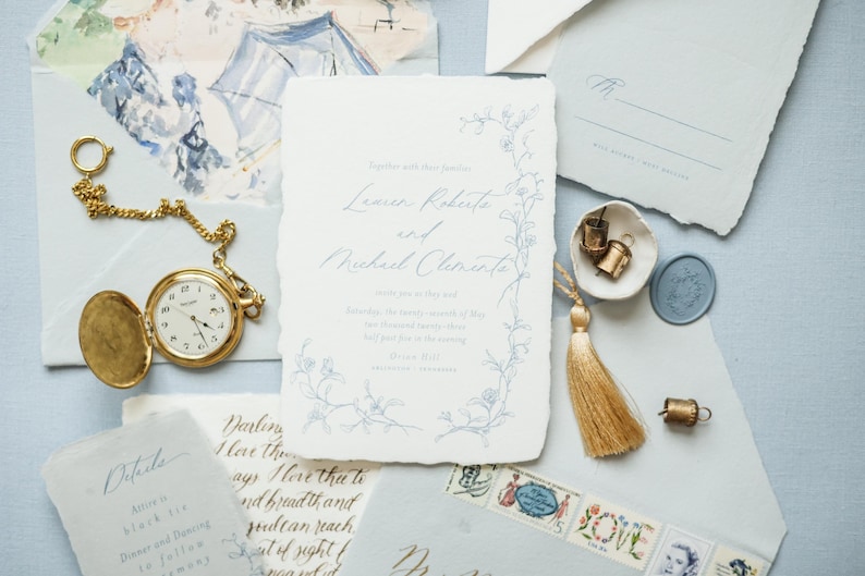 May include: A wedding invitation suite with a delicate floral design in blue and white. The invitation features the names "Lexus Rokute" and "Michiel Clemente" in elegant calligraphy. The suite includes a pocketfold envelope, a RSVP card, and a wax seal.