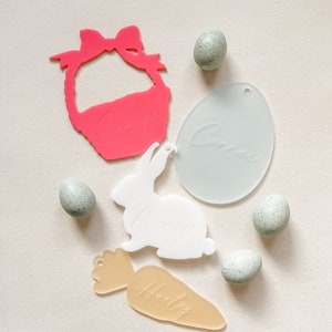 Puede incluir: Tres decoraciones de Pascua de acrílico con nombres escritos en ellas. Una cesta rosa con el nombre "Grayson", un conejito blanco con el nombre "Avery" y una zanahoria tostada con el nombre "Hailey".