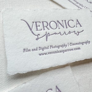 May include: Close-up of several white business cards with torn edges. The cards feature the name "VERONICA sparrow" in a purple script font, along with the text "Film and Digital Photography | Cinematography" and a website address.