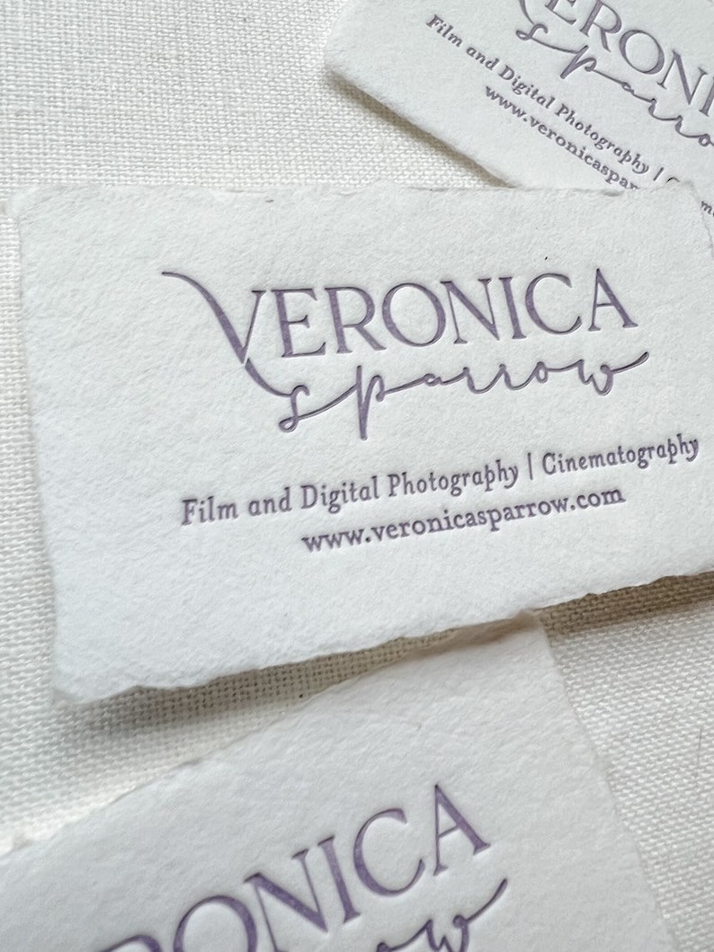 May include: Close-up of several white business cards with torn edges. The cards feature the name "VERONICA sparrow" in a purple script font, along with the text "Film and Digital Photography | Cinematography" and a website address.