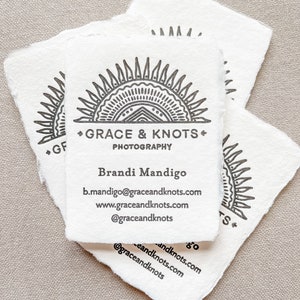 May include: Several white business cards with a deckled edge are stacked. The cards feature a black sunburst design with the text "GRACE & KNOTS PHOTOGRAPHY" and contact information for Brandi Mandigo.