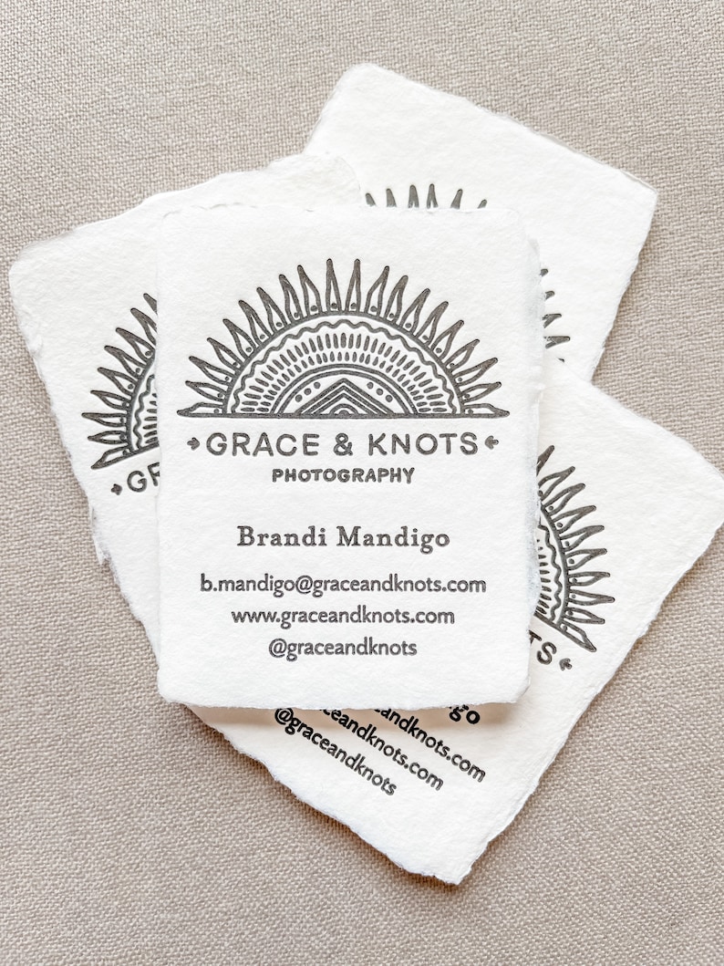 May include: Several white business cards with a deckled edge are stacked. The cards feature a black sunburst design with the text "GRACE & KNOTS PHOTOGRAPHY" and contact information for Brandi Mandigo.