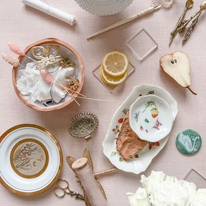 May include: A flat lay of various craft supplies on a pink background. The items include a white bowl filled with buttons, a white plate with a gold design, a spool of thread, a pair of scissors, a small white bowl with a butterfly charm, a small white plate with a strawberry design, a pear, a lemon, and a bouquet of white roses.