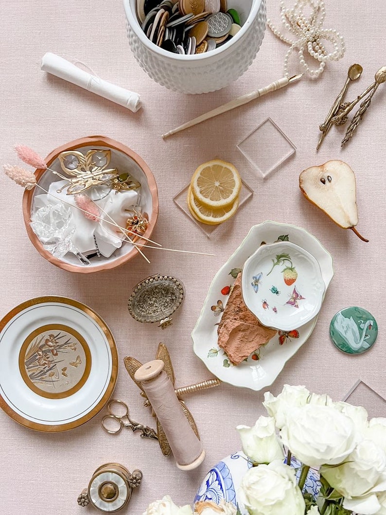 May include: A flat lay of various craft supplies on a pink background. The items include a white bowl filled with buttons, a white plate with a gold design, a spool of thread, a pair of scissors, a small white bowl with a butterfly charm, a small white plate with a strawberry design, a pear, a lemon, and a bouquet of white roses.