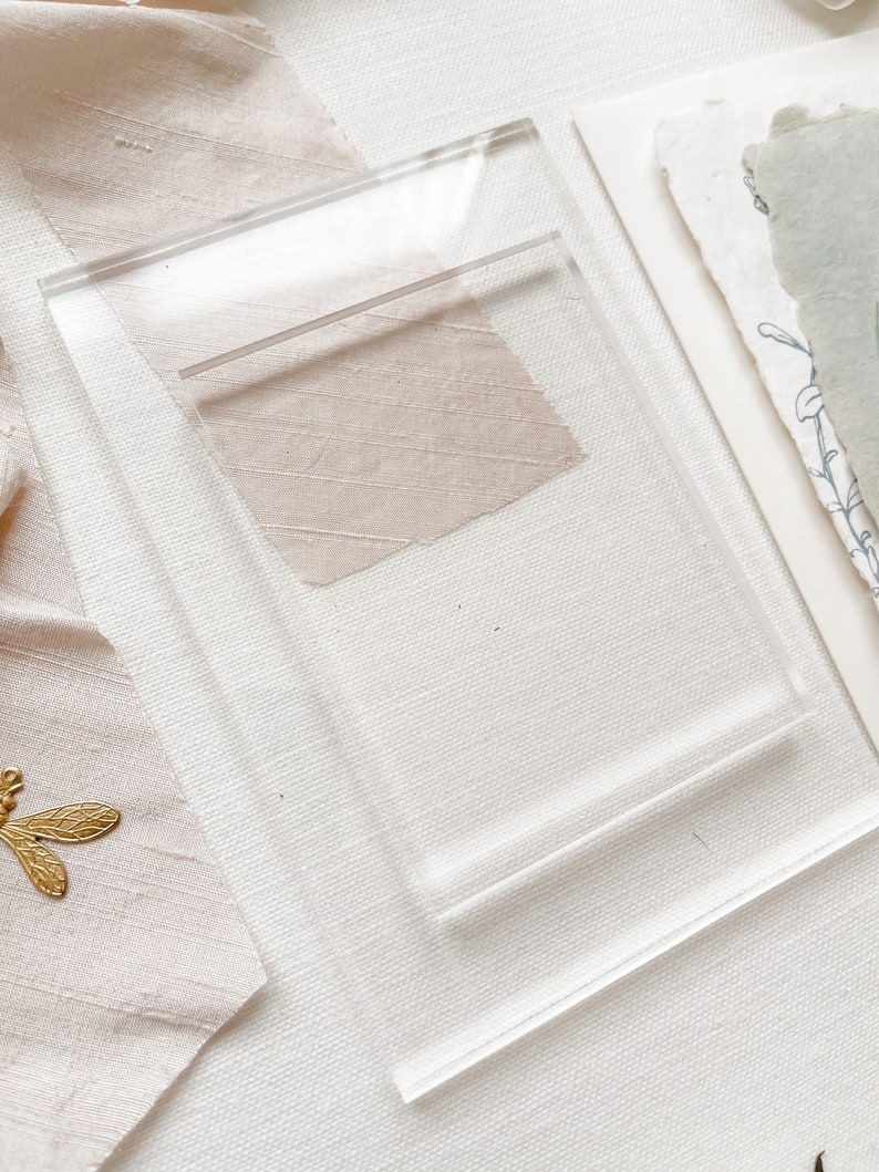 May include: A clear acrylic display stand with a raised platform. The stand is designed to hold and display small items, such as jewellery, cards, or other decorative objects.
