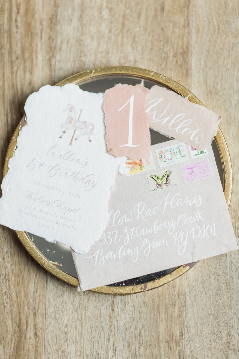 May include: A white card with the text "Willow's 1st Birthday" and the date "December 1, 2015". The card also includes the text "Taylors Chapel" and an address. The card is on a gold tray with a pink card with the number "1" and a white card with the text "Willow".