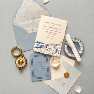 May include: A wedding invitation suite with a blue and white color scheme. The invitation features a hand-drawn illustration of a cityscape. The text on the invitation reads "Tiffany Dawn Hays and Lewis Winston Silvers request the pleasure of your company as they wed... Tuesday, the fifth of March... Thira".