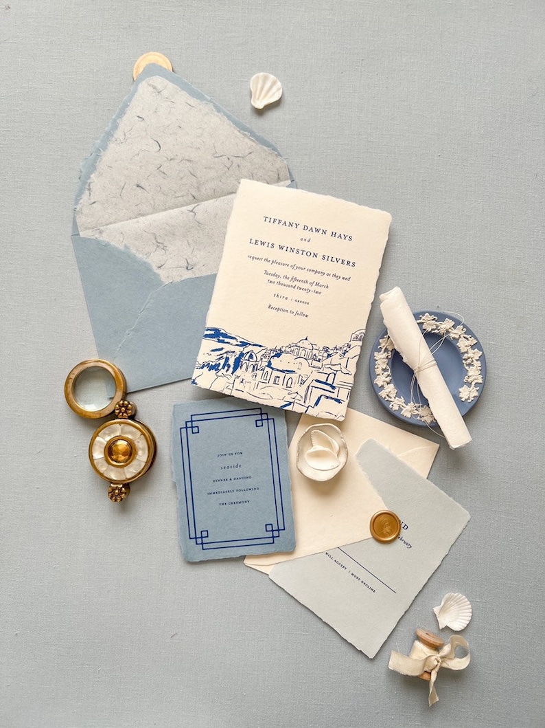 May include: A wedding invitation suite with a blue and white color scheme. The invitation features a hand-drawn illustration of a cityscape. The text on the invitation reads "Tiffany Dawn Hays and Lewis Winston Silvers request the pleasure of your company as they wed... Tuesday, the fifth of March... Thira".