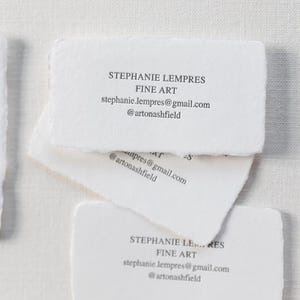 May include: A stack of white business cards with torn edges. The cards display the name "STEPHANIE LEMPRES" and "FINE ART", along with an email address and social media handle. The cards are arranged on a white surface.