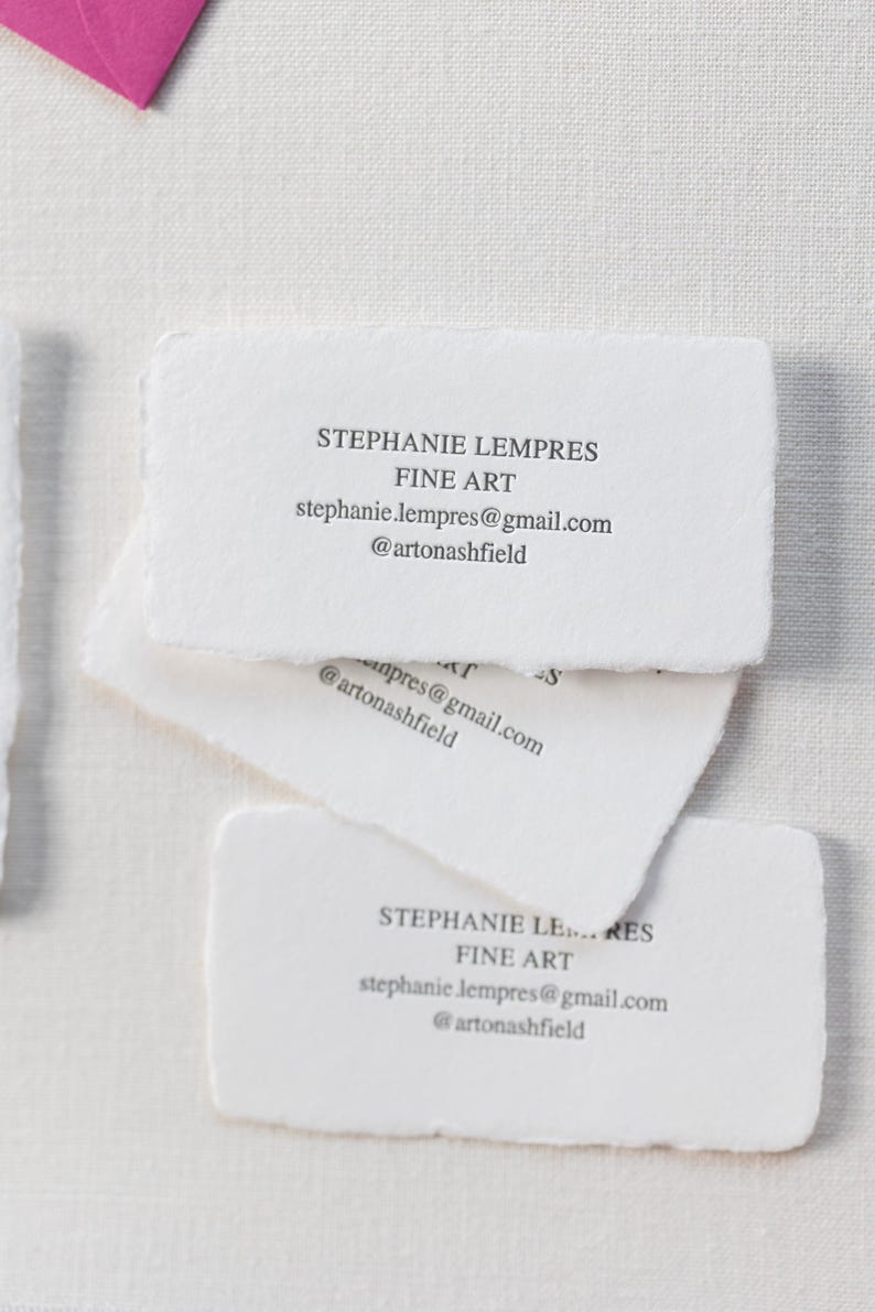 May include: A stack of white business cards with torn edges. The cards display the name "STEPHANIE LEMPRES" and "FINE ART", along with an email address and social media handle. The cards are arranged on a white surface.