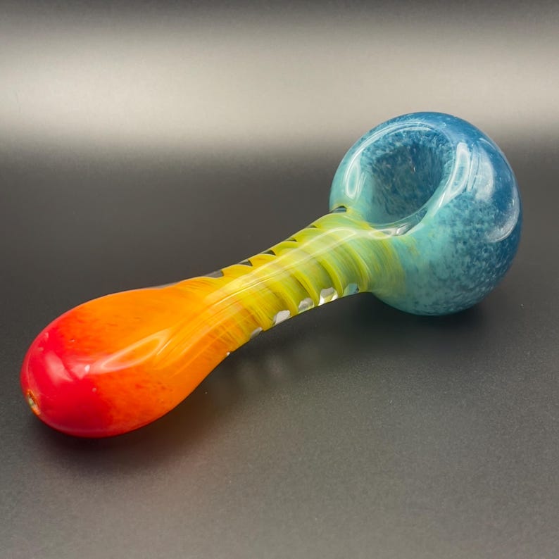 Op de afbeelding: Een glazen pijp met een kleurovergang van rood naar geel en blauw. De kop is blauw, de steel geel en oranje. De pijp heeft een afgeronde vorm en is bedoeld om te roken.