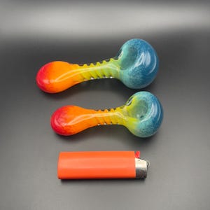 Op de afbeelding: Twee glazen pijpen met een kleurengradatie, waaronder rood, oranje, geel en blauw. De pijpen liggen naast een oranje aansteker op een donkere ondergrond.