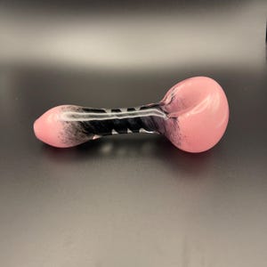 Op de afbeelding: Een roze en zwarte glazen pijp met een afgeronde kom en een cilindrisch lichaam. De pijp heeft een roze tint aan de uiteinden, die overgaat in zwart met witte accenten in het midden. Het ontwerp is glad en lijkt handgemaakt.