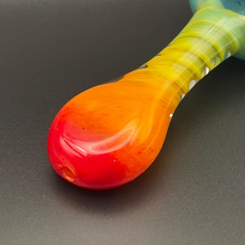 Op de afbeelding: Een kleurrijke glazen pijp met een kleurengradi&euml;nt. De kom is rood, overgaand in oranje, dan geel en tenslotte groen. De pijp heeft een gladde, glanzende afwerking en een klein gaatje aan het einde van de kom.