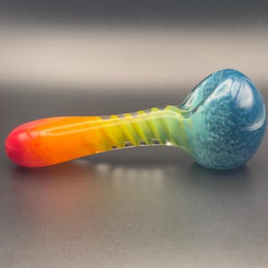 Op de afbeelding: Een kleurrijke glazen pijp met een kom en steel. De kom is gespikkeld blauw, gaat over in een groene en gele spiraal en eindigt met een rood en oranje mondstuk. De pijp is ontworpen om te roken.