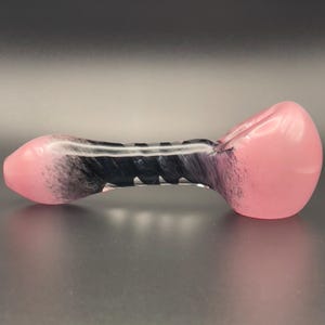 Op de afbeelding: Een roze en zwarte glazen pijp met een afgeronde kom en mondstuk. De pijp heeft een zwart-wit wervelontwerp in het midden. Het glas is glad en lijkt mondgeblazen.