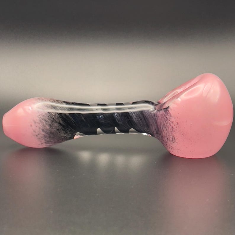 Op de afbeelding: Een roze en zwarte glazen pijp met een afgeronde kom en mondstuk. De pijp heeft een zwart-wit wervelontwerp in het midden. Het glas is glad en lijkt mondgeblazen.