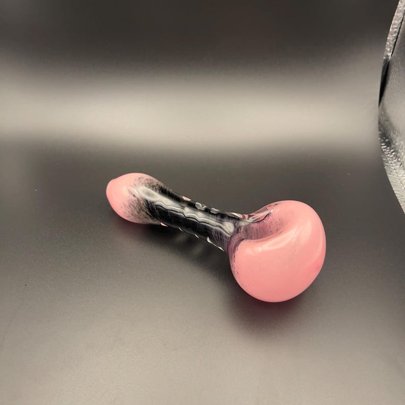 Op de afbeelding: Een roze en zwarte glazen pijp met een afgeronde kom en een cilindrische steel. De pijp heeft een gladde, glanzende afwerking met een wervelpatroon van roze en zwart. De steel heeft een reeks kleine, verhoogde bobbels.