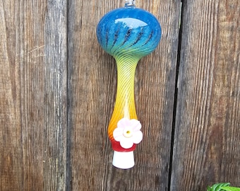 Könnte beinhalten: Ein farbenfrohes Windspiel aus Glas mit blauem und gelbem Design, verziert mit einer kleinen rosa Blume. Das Windspiel hängt an einer silbernen Kette vor einem Holzgrund.