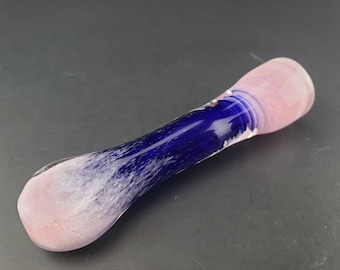 May include: A glass pipe with a pink and blue design. The mouthpiece and bowl are pink, while the center is a deep blue. The colors blend together, creating a unique aesthetic. The pipe is smooth and appears to be handmade.