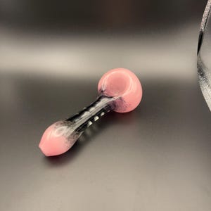 Op de afbeelding: Een roze en zwarte glazen pijp met een afgeronde kom en een lange, slanke steel. De kom en het mondstuk zijn effen roze, terwijl de steel een zwart-wit ontwerp heeft. De pijp is ontworpen om te roken.