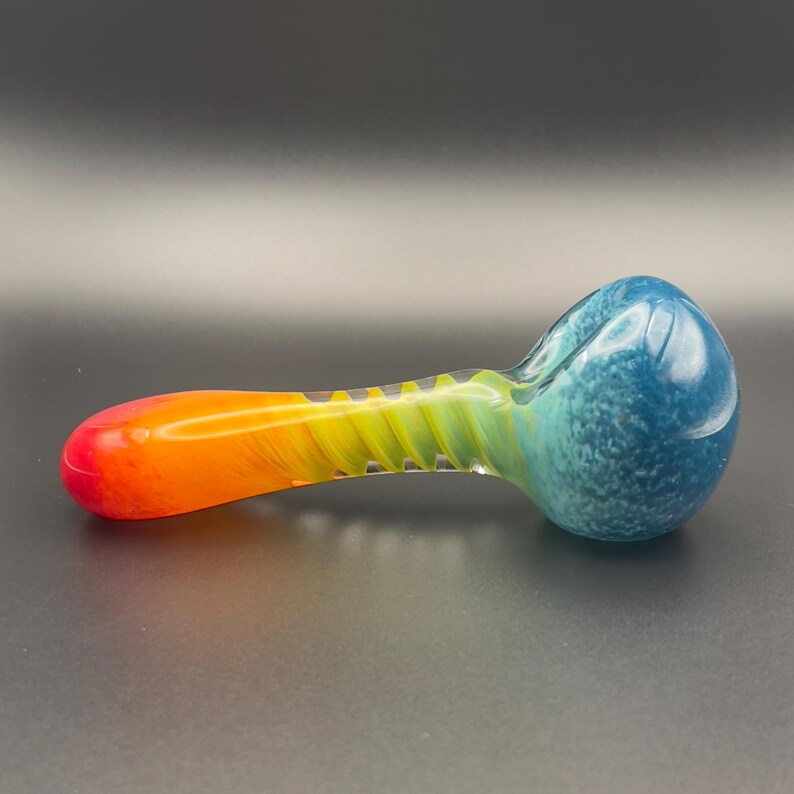 Op de afbeelding: Een kleurrijke glazen pijp met een regenboogverloop. De kom is diepblauw met witte spikkels, gaat over in geel en oranje en eindigt in een rode punt. De steel heeft een spiraalvormig ontwerp.