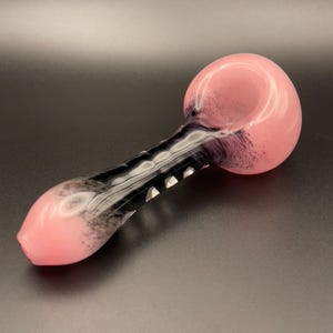 Op de afbeelding: Een roze en zwarte glazen pijp met een afgeronde kom en een lange, gebogen steel. De kom en het mondstuk zijn zachtroze van kleur, terwijl de steel een zwart-wit wervelontwerp heeft. De pijp is ontworpen om te roken.